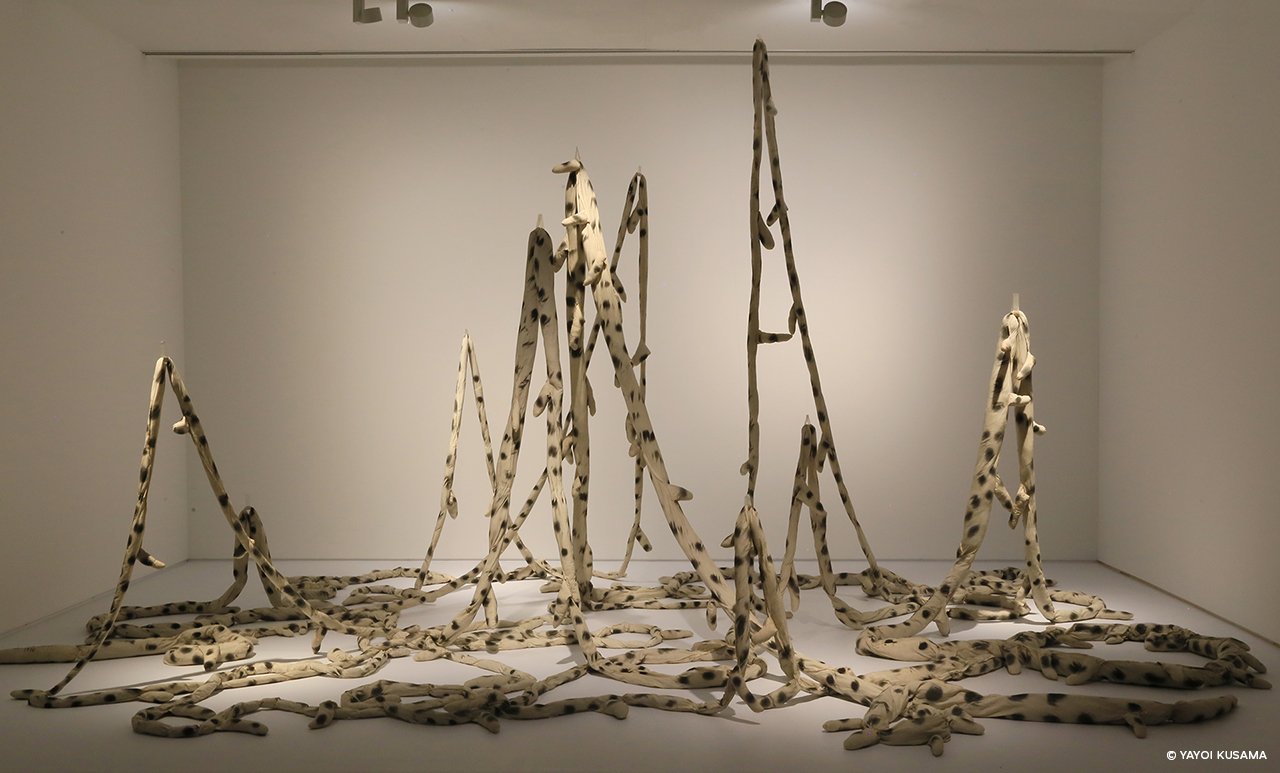 Installation artwork in which an elongated piece of stuffed fabric dangles at various hanging points and cluster in a loose muddle on the floor.